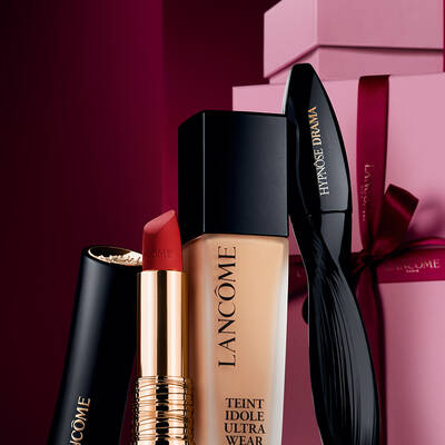 Ein elegantes Stillleben mit dem matten L'Absolu Rouge Lippenstift, der Teint Idole Ultra Wear Foundation und der Hypnose Drama Mascara von Lancome vor einem luxuriosen, dunkelroten Weihnachtshintergrund.