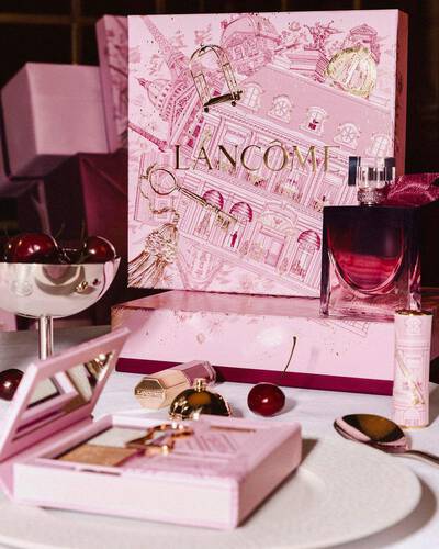 Abbildung der festlichen Lancome-Weihnachtskollektion auf einem Tisch, bestehend aus einer großen, illustrierten rosa Geschenkbox, Parfum, Make-up-Produkten und frischen Kirschen.