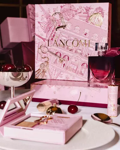 Abbildung der festlichen Lancome-Weihnachtskollektion auf einem Tisch, bestehend aus einer großen, illustrierten rosa Geschenkbox, Parfum, Make-up-Produkten und frischen Kirschen.