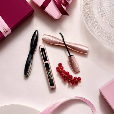 Eine flache Anordnung von Lancome-Make-up-Produkten, darunter Mascara und Lipgloss, zusammen mit rosa Geschenkboxen, Satinbandern und einem Zweig roter Johannisbeeren.