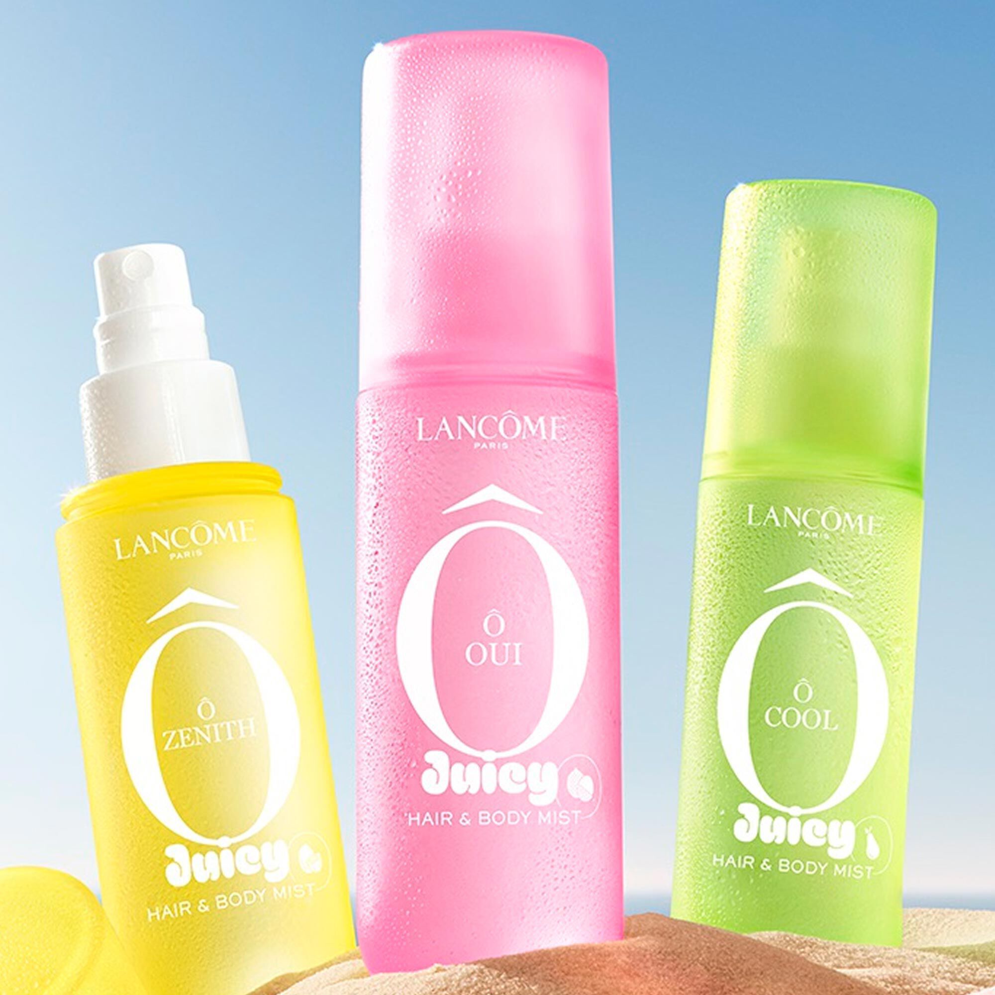 Drei bunte Spruhflaschen, eine gelb, eine rosa und eine grun, stehen an einem Sandstrand neben einem rosa gestreiften Handtuch. Der Ozean ist im Hintergrund unter klarem blauen Himmel zu sehen.