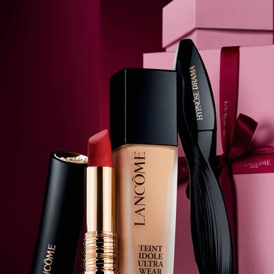 Ein elegantes Stillleben mit dem matten L'Absolu Rouge Lippenstift, der Teint Idole Ultra Wear Foundation und der Hypnose Drama Mascara von Lancome vor einem luxuriosen, dunkelroten Weihnachtshintergrund.