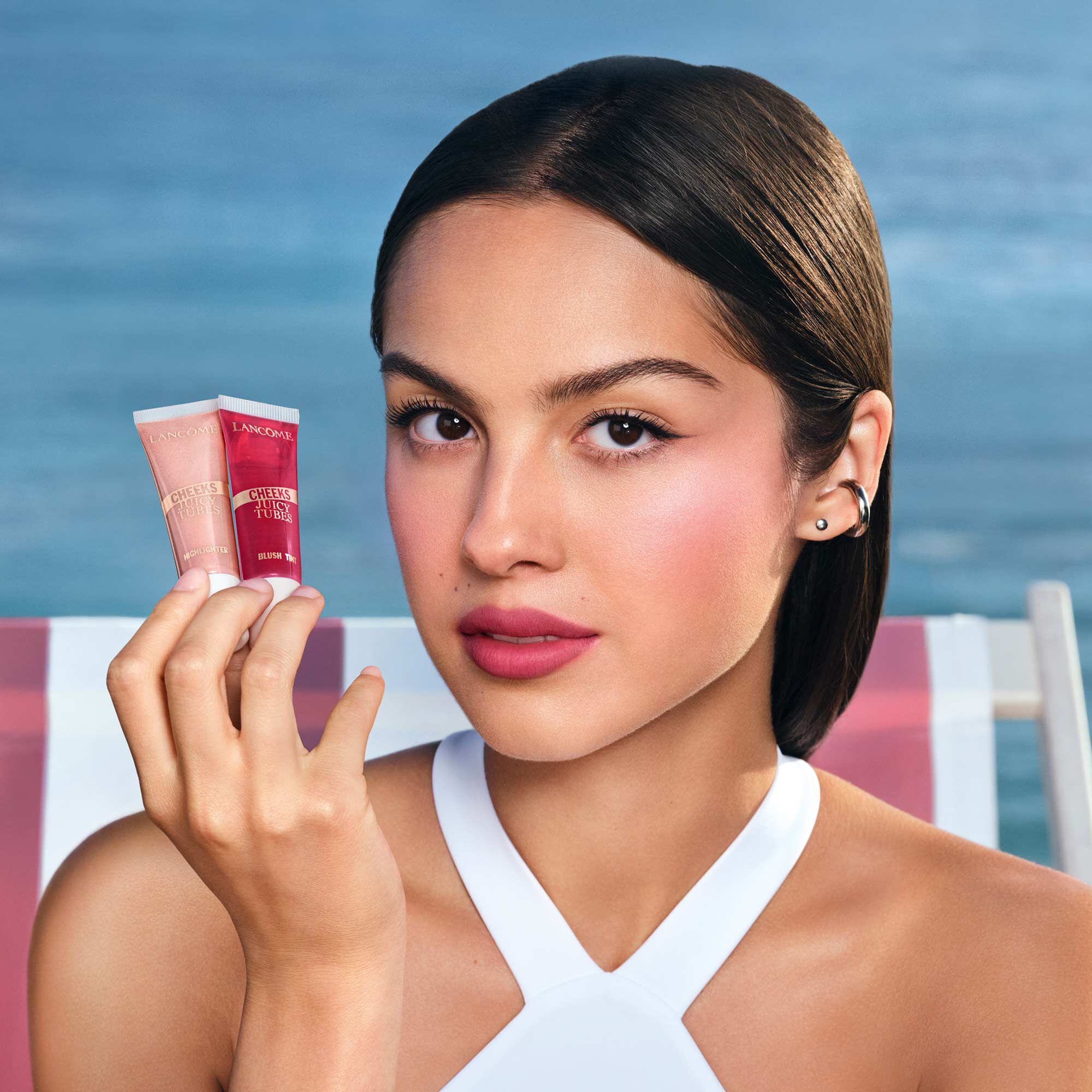 Olivia Rodrigo halt einen Juicy Tubes Highlighter mit Strandhintergrund.