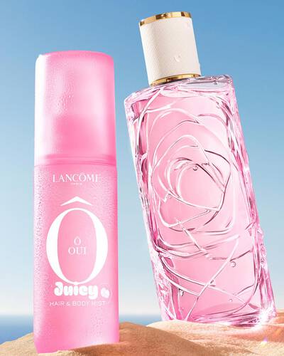 Drei bunte Spruhflaschen, eine gelb, eine rosa und eine grun, stehen an einem Sandstrand neben einem rosa gestreiften Handtuch. Der Ozean ist im Hintergrund unter klarem blauen Himmel zu sehen.
