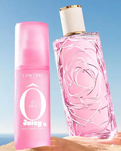 Drei bunte Spruhflaschen, eine gelb, eine rosa und eine grun, stehen an einem Sandstrand neben einem rosa gestreiften Handtuch. Der Ozean ist im Hintergrund unter klarem blauen Himmel zu sehen.