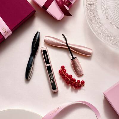 Eine flache Anordnung von Lancome-Make-up-Produkten, darunter Mascara und Lipgloss, zusammen mit rosa Geschenkboxen, Satinbandern und einem Zweig roter Johannisbeeren.
