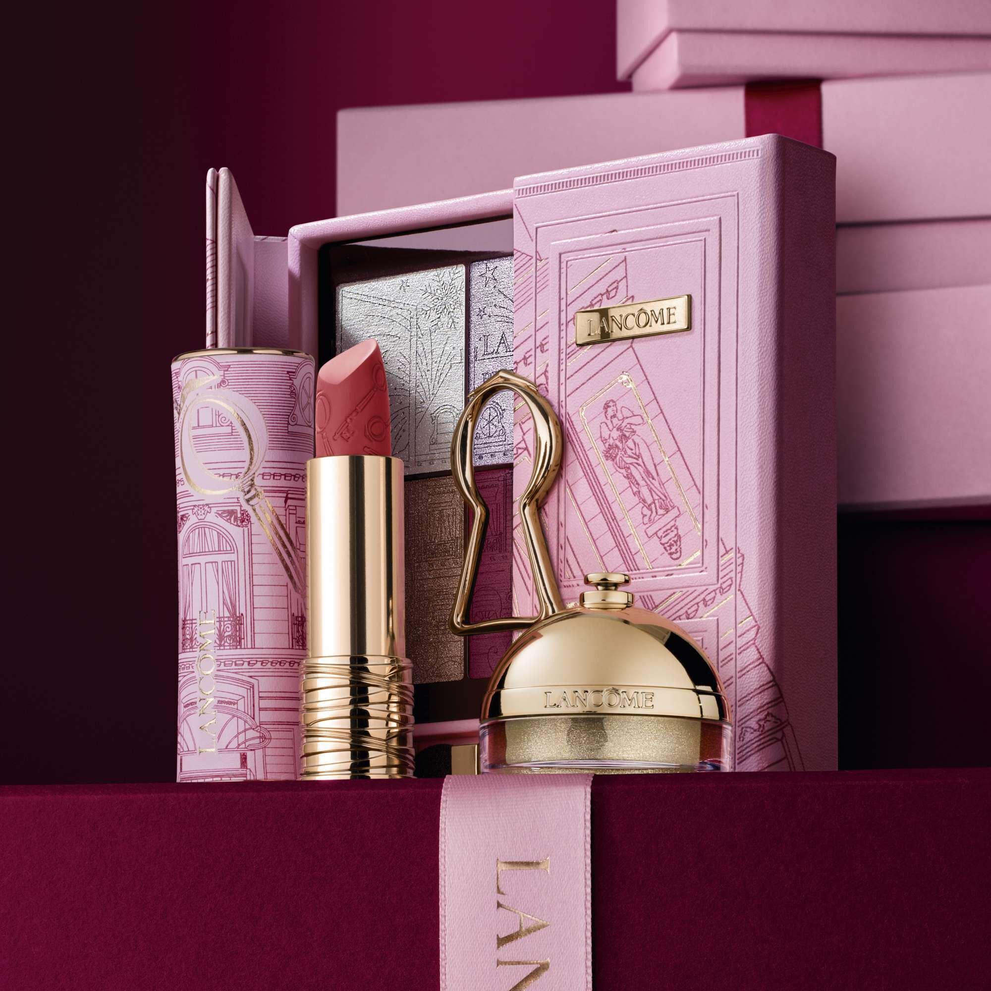 Abbildung der Drama Matte, Ring & Shine und der The Suite Escape Limited Edition Beauty-Essentials vor einem Stapel rosa und dunkelroter Geschenkboxen vor einem dunkelroten Hintergrund.