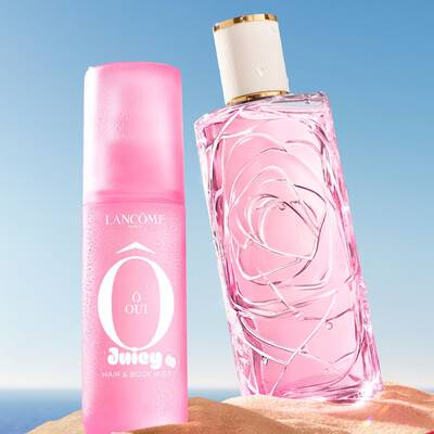 Eine Strandszene mit drei bunten Parfumflaschen, die aufrecht im Sand stehen, eine pinke, eine gelbe und eine grune, mit einer weiteren rosa Parfumflasche, die im Vordergrund auf der Seite liegt. Ein rosa-weiß gestreiftes Handtuch liegt links uber dem Sand. Der Hintergrund zeigt einen klarblauen Himmel, der am Horizont in einen blauen Ozean ubergeht.