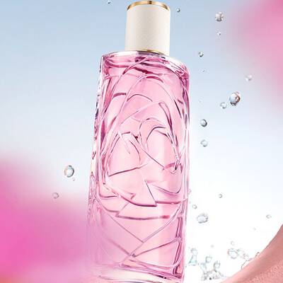 Eine hellrosa Parfumflasche mit strukturiertem Glasdesign vor einem hellblauen Himmel, um den Wassertropfen herum spritzten. Die Flasche ist durch eine runde Offnung in einer strukturierten rosa Wand zu sehen, und im Vorder- und Hintergrund befinden sich verschwommene rosa Blumen.