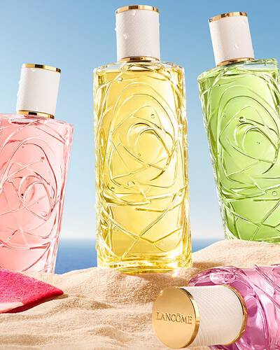 Zwei rosa Flaschen, eine parfumtransparente Glasflasche mit strukturiertem Design und eine Spruhflasche, die an einem Sandstrand steht. Ein gefaltetes, rosa gestreiftes Strandhandtuch liegt neben den Flaschen. Im Hintergrund gibt es einen klarblauen Himmel und das Meer.