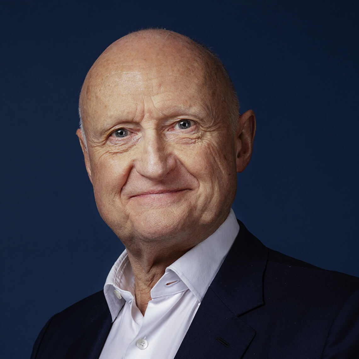 Portrat eines alteren Mannes mit Glatze und freundlich lachelndem Gesicht, vor dunklem Hintergrund.