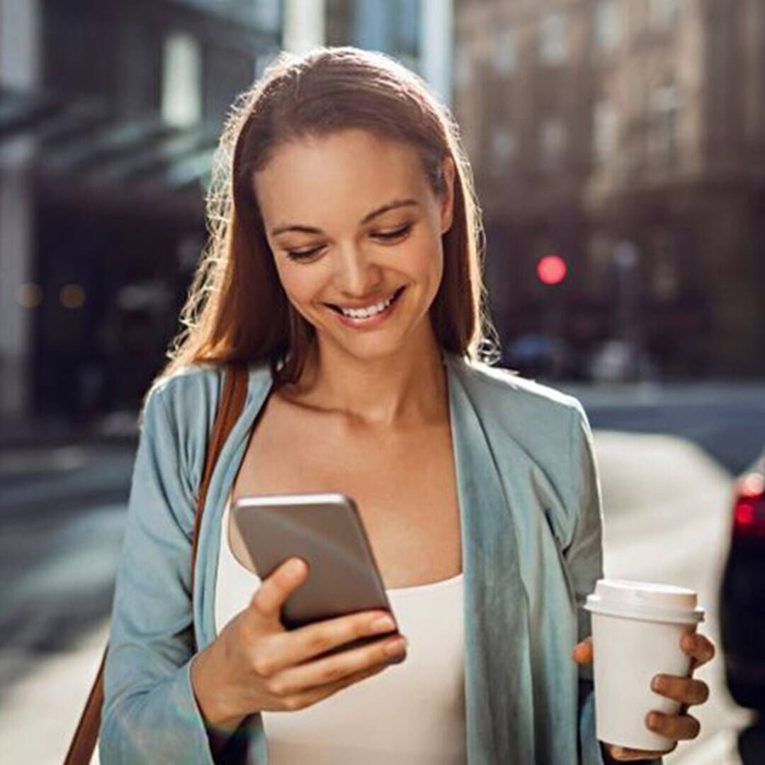 Eine Frau lauft uber die Straße und schaut lachend auf ihr Handy, in der anderen Hand halt sie einen Kaffeebecher.