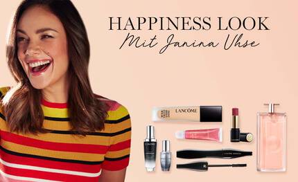 Eine lachelnde Frau mit Make-up-Produkten und einem Parfum im Hintergrund, begleitet von der Aufschrift "Happiness Look mit Janina Uhe".
