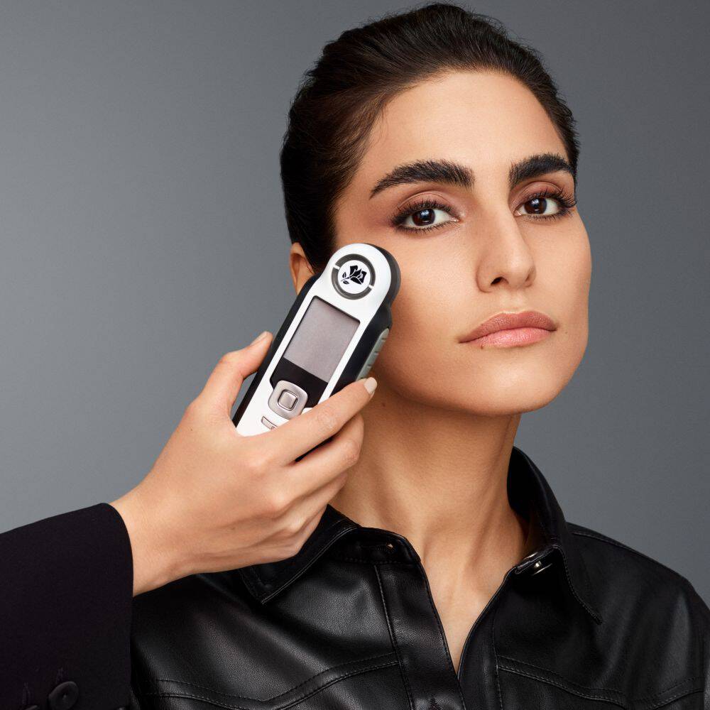 Nahaufnahme einer Frau mit dunklem Haar und schwarzer Lederjacke, an deren Wange ein tragbares Lancome-Hautanalysegerat (Shade Finder) gehalten wird, vor einem neutralen grauen Hintergrund.