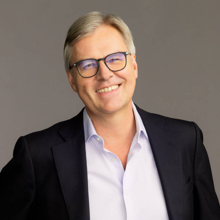Ein lachelnder Mann mit Brille, in einem dunklen Blazer und hellen Hemd, vor einem grauen Hintergrund.