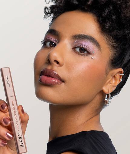 Eine junge Frau mit lockigem Haar, einem strahlenden Teint und glitzerndem Augenmake-up halt einen Mascara von Lancome in der Hand.