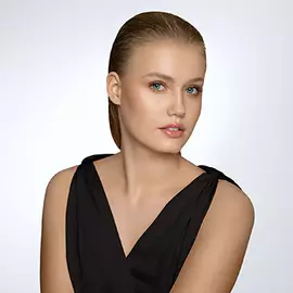 Eine Frau mit glattem, zuruckgegeltem Haar und naturlichem Make-up tragt ein schwarzes Oberteil. Sie blickt direkt in die Kamera und hat eine neutrale Hintergrundbeleuchtung.