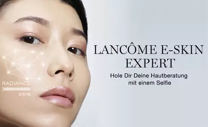 Eine Frau mit glatter Haut betrachtet sich im Spiegel, wahrend sie auf ein digitales Hautanalyse-Tool auf ihrem Gesicht schaut. Der Text sagt: "LANCOME E-SKIN EXPERT - Hole Dir Deine Hautberatung mit einem Selfie."