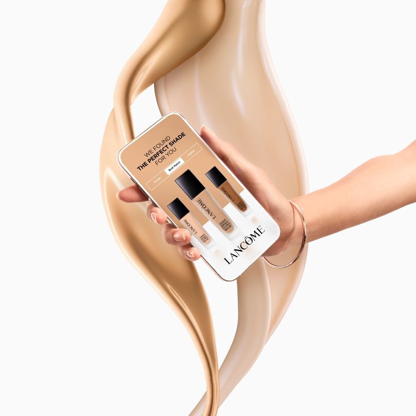 Hand halt Smartphone mit Lancome Foundation-Farbabgleich vor wirbelndem Foundation-Hintergrund.
