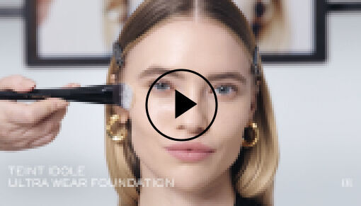 Eine Nahaufnahme einer Person, die mit einem Pinsel eine ubertragbare Foundation auftragt, wahrend ein Video-Playback-Symbol im Vordergrund angezeigt wird.