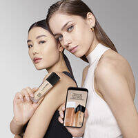 Zwei Model prasentieren eine Foundation-Flasche und ein Smartphone mit Produktinfos, vor einem grauen Hintergrund.