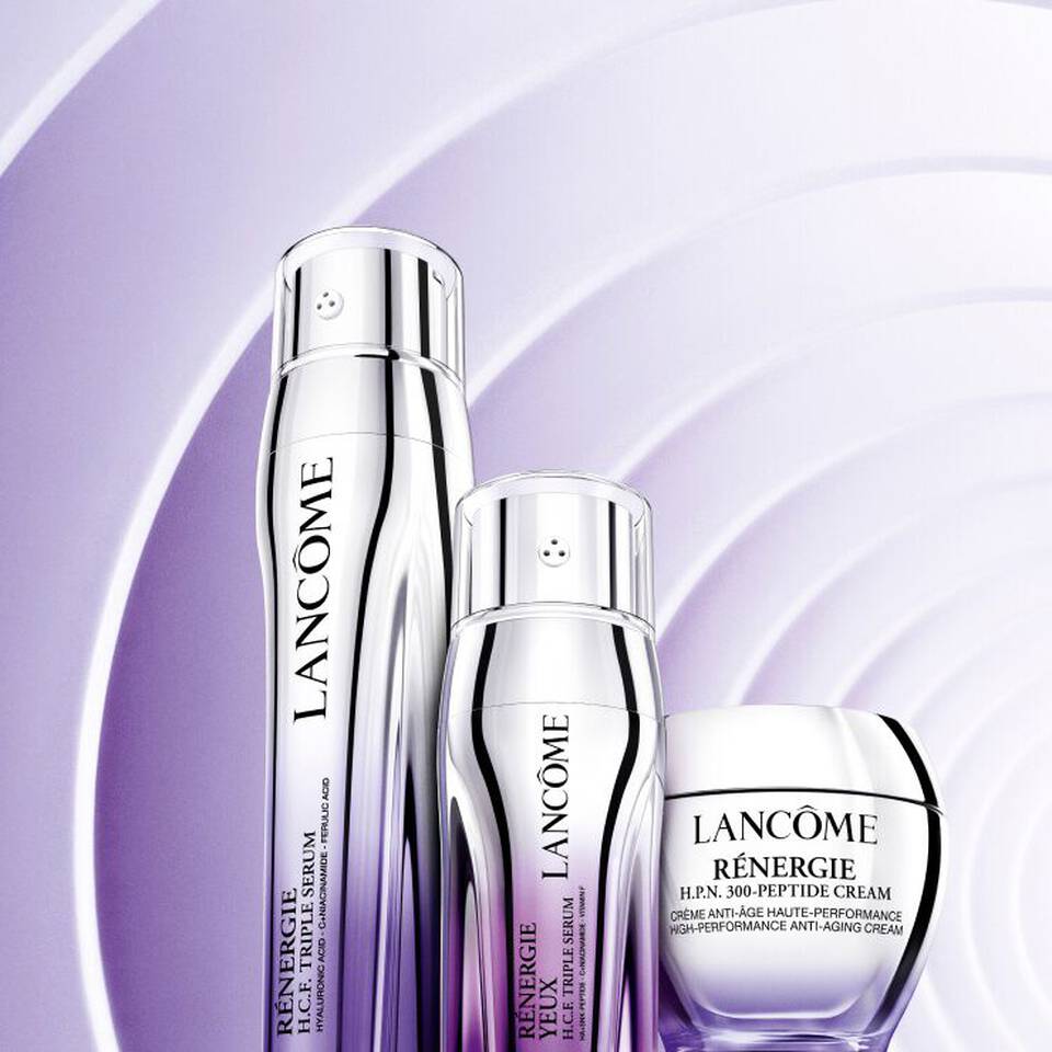 Drei Lancome Hautpflegeprodukte in eleganter Verpackung vor lila Wellenhintergrund. Anti-Aging und Hyaluronsaure betont.