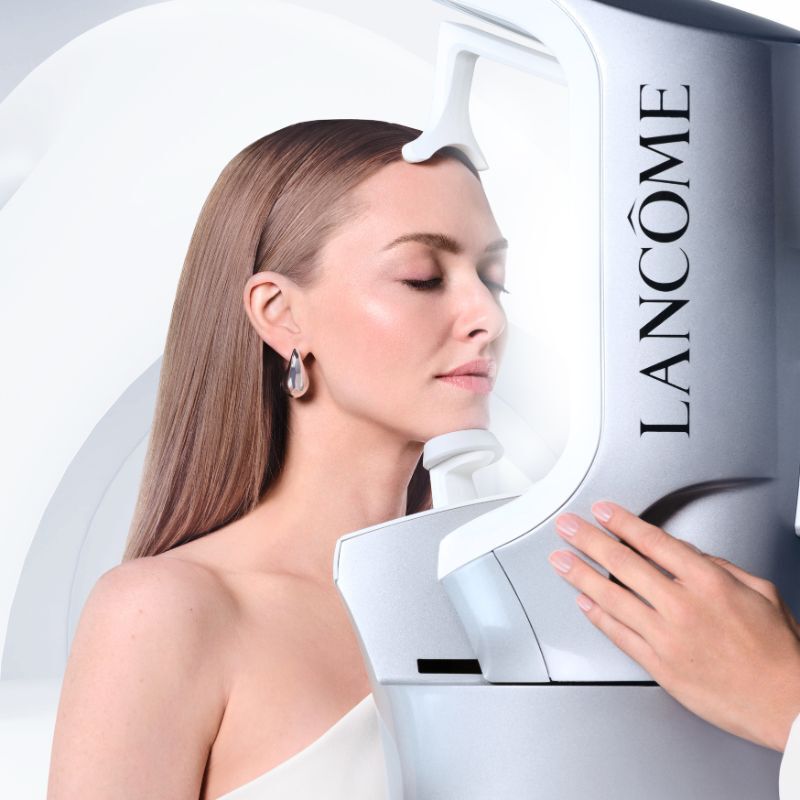 Amanda Seyfried mit geschlossenen Augen bei einer professionellen Hautanalyse mit dem Lancome Skin Screen Gerat vor einem geschwungenen, weißen abstrakten Hintergrund.