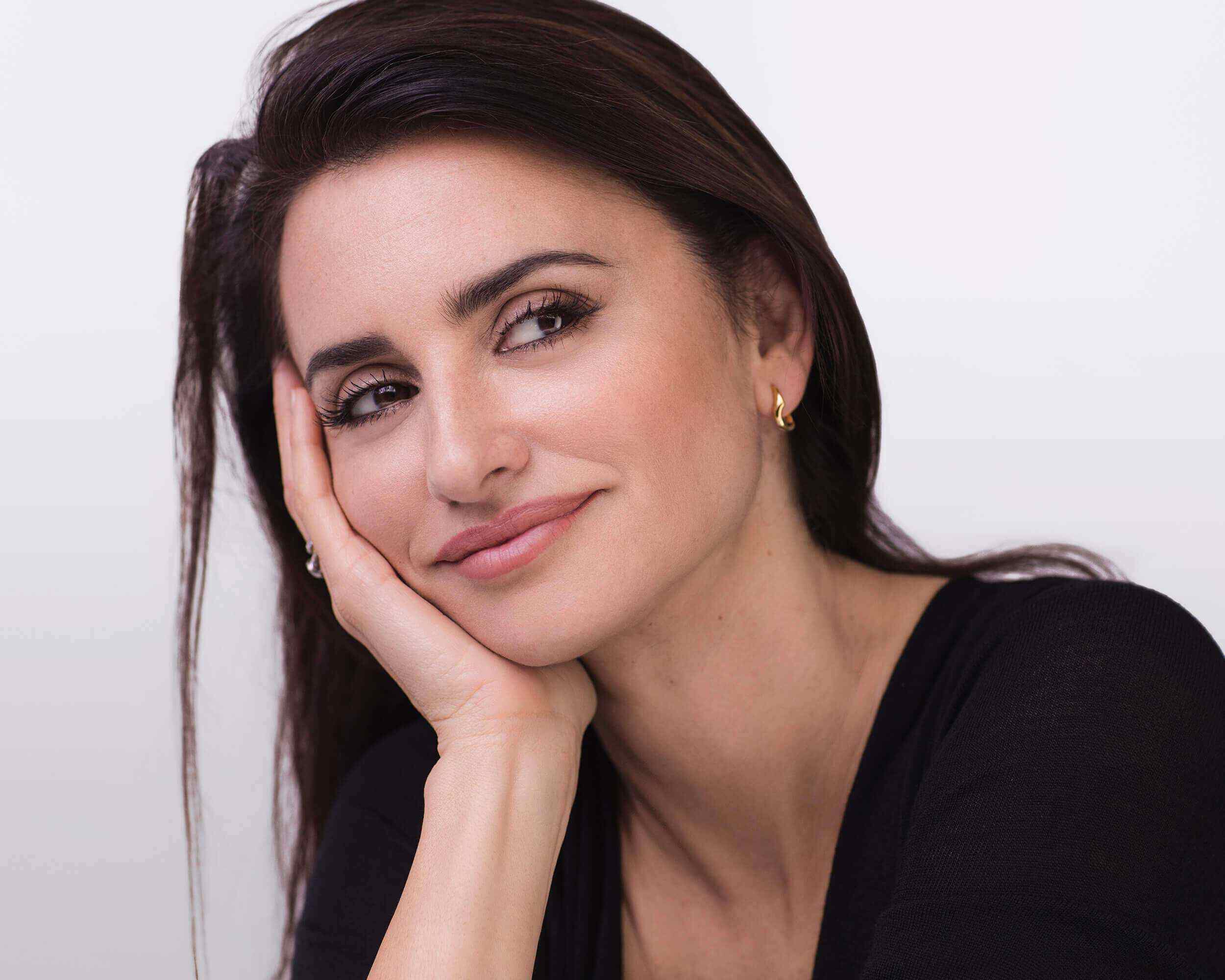 Eine Frau mit langen, dunklen Haaren lachelt sanft, wahrend sie ihren Kopf auf die Hand stutzt. Sie tragt ein schwarzes Oberteil und hat einen freundlichen Ausdruck.