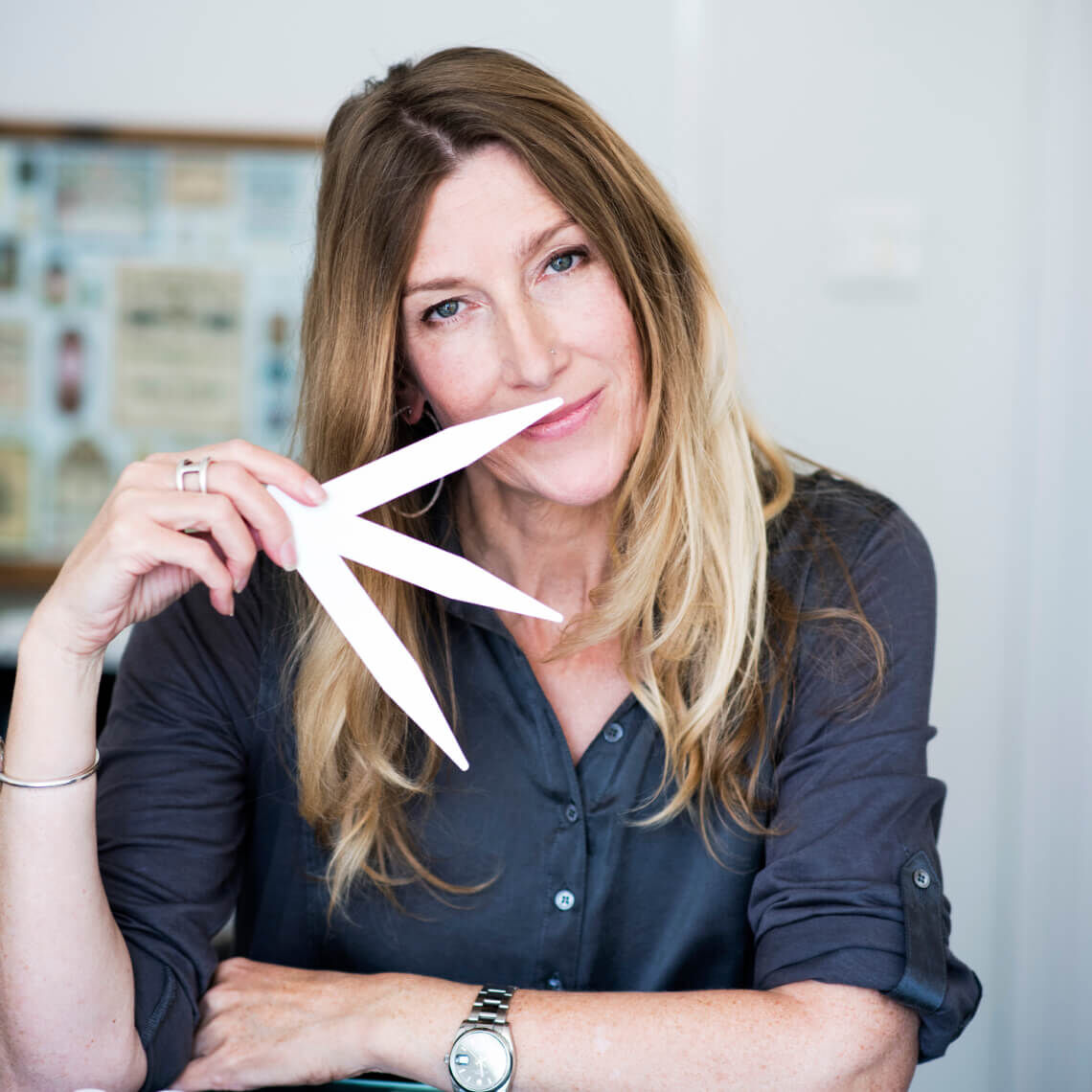 Eine Frau mit langen, blonden Haaren halt ein weißes Papierobjekt vor ihrem Gesicht und lachelt freundlich.