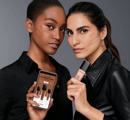 Zwei Frauen prasentieren verschiedene Make-up-Produkte und halten ein Smartphone in der Hand, auf dem die passende Foundationfarbe angezeigt wird.