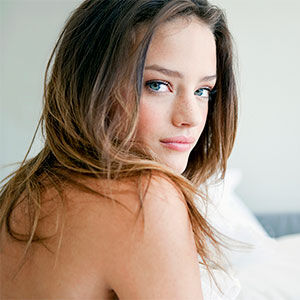 Eine Frau sitzt Oberkorperfrei im Bett und schaut uber die Schulter in Richtung Kamera.