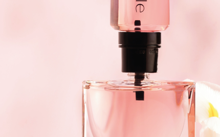 Ein Parfumflakon mit rosafarbenem Inhalt steht auf einem rosa Hintergrund, umgeben von Pflanzen.
