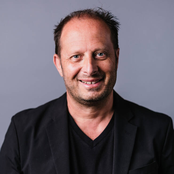Lachelnder Mann in schwarzem Sakko vor neutralem Hintergrund. Freundlicher, selbstbewusster Gesichtsausdruck.