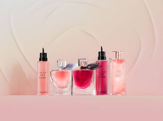 Zwei Hande, eine behandschuht und eine unhandschuht, interagieren mit zwei Lancome "La Vie Est Belle" Parfumflaschen vor einer Berg- und Himmelskulisse.