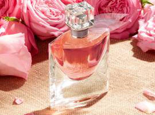 Zwei Hande, eine behandschuht und eine unhandschuht, interagieren mit zwei Lancome "La Vie Est Belle" Parfumflaschen vor einer Berg- und Himmelskulisse.