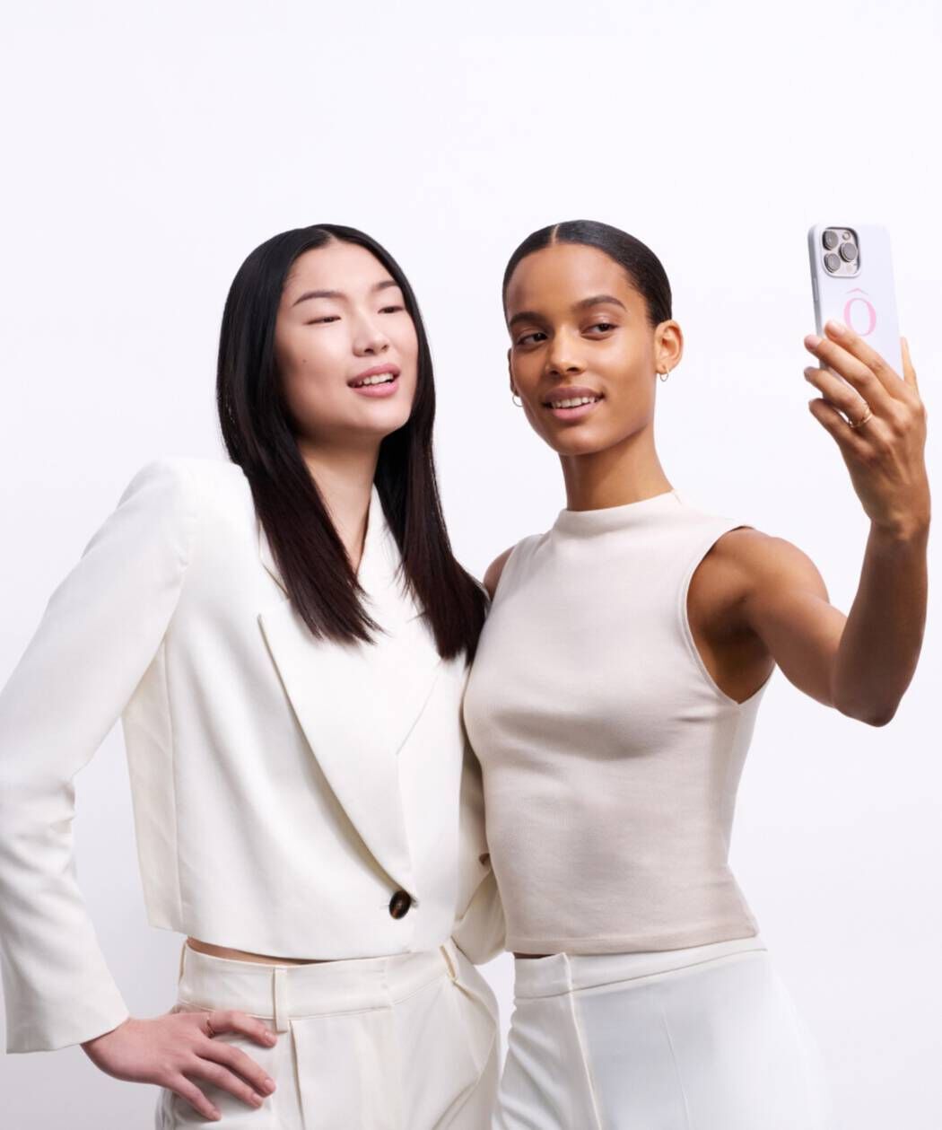 Zwei Frauen stehen nebeneinander und machen ein Selfie mit einem Smartphone. Beide tragen helle, elegante Kleidung und lacheln.