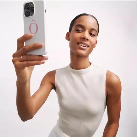 Eine junge Frau halt ein Smartphone in der Hand und macht ein Selfie. Sie tragt ein helles, armelloses Oberteil und hat einen lachelnden Gesichtsausdruck.