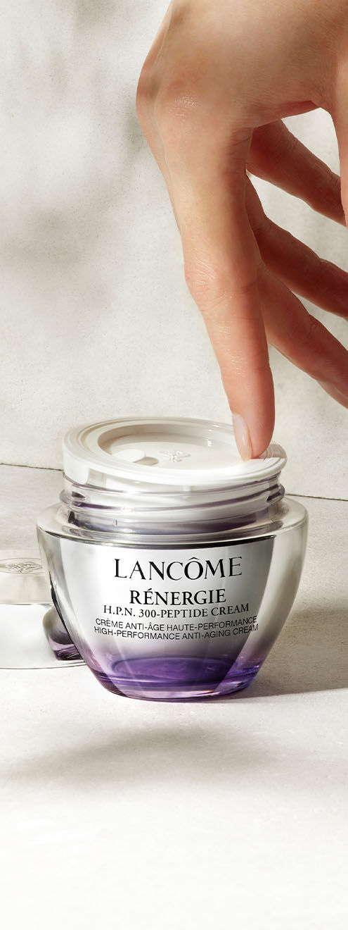 Eine Hand greift nach einem Glastiegel mit Lancome Renergie Anti-Aging-Peptidcreme auf einem hellen, strukturierten Hintergrund.