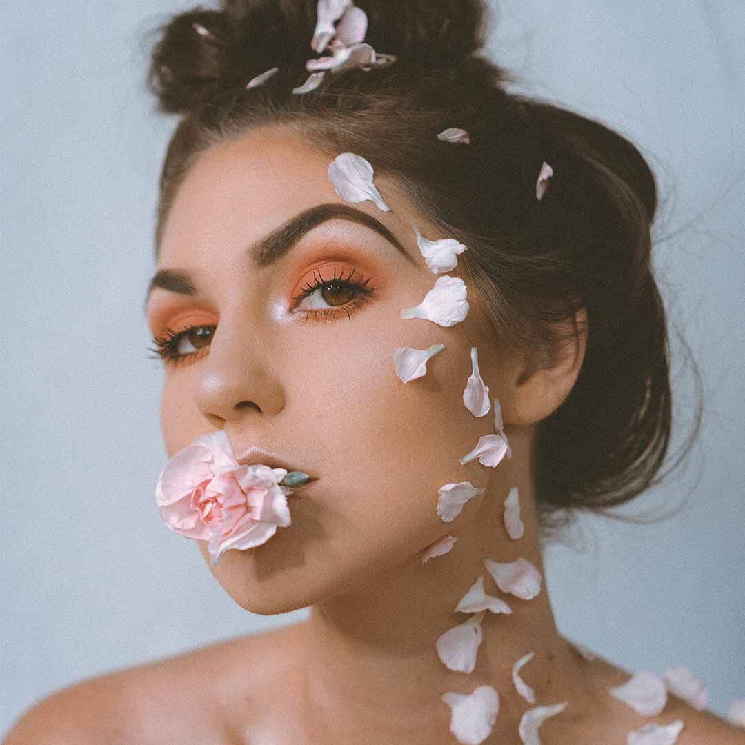 Eine Frau mit einer rosa Blume im Mund und Blutenblattern im Gesicht, geschminkt mit orangefarbenem Lidschatten.