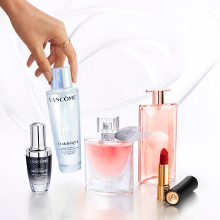 Eine Hand greift nach einem Hautpflegeprodukt von Lancome, umgeben von weiteren Lancome-Produkten wie Serum, Parfum und Lippenstift auf einem hellen Hintergrund.