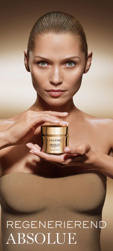 Eine Frau mit zuruckgebundenem Haar und grunen Augen halt einen goldenen Tiegel Lancome Absolue Creme und blickt direkt in die Kamera. Unten steht der Text „REGENERIEREND ABSOLUE“.