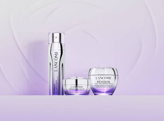Drei Lancome Renergie Produkte – eine Pumpflasche, ein Tiegel Augencreme und ein Tiegel Gesichtscreme – auf einem hellvioletten Hintergrund mit sanften Wirbelmustern.