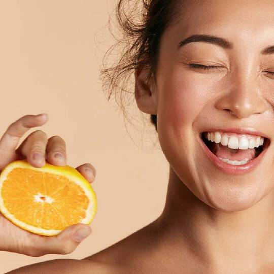 Eine lachelnde Frau mit geschlossenen Augen halt eine frische Orangenscheibe vor einem neutralen Hintergrund.