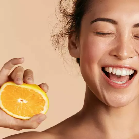 Eine lachelnde Frau mit geschlossenen Augen halt eine frische Orangenscheibe vor einem neutralen Hintergrund.