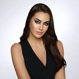 Portrat einer Frau mit langen, braunen Haaren und auffalligem Make-up, das grune und blaue Tone enthalt. Sie tragt ein schwarzes Oberteil und hat einen neutralen Gesichtsausdruck vor einem hellen Hintergrund.