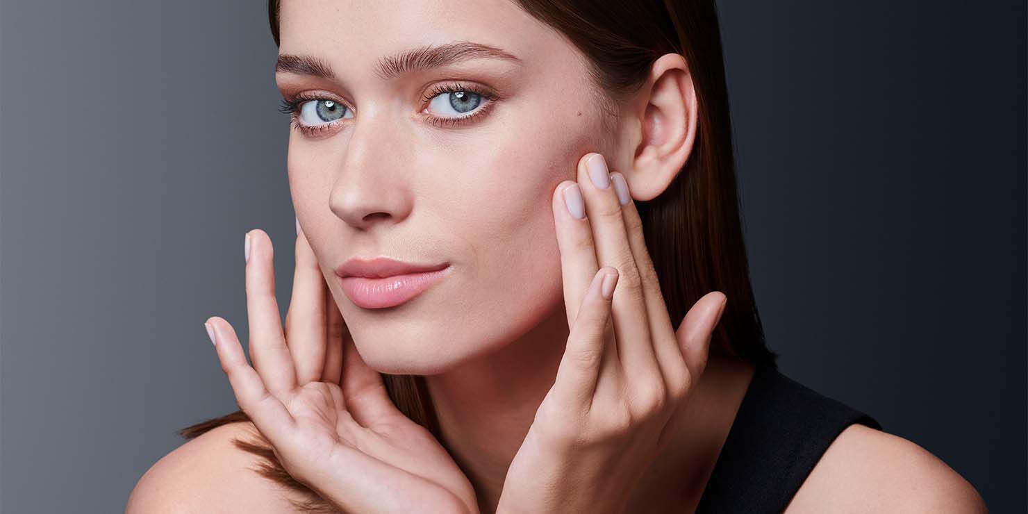 Eine Frau mit naturlichem Make-up und hellrosa Lippen beruhrt sanft ihr Gesicht mit beiden Handen und blickt direkt in die Kamera.