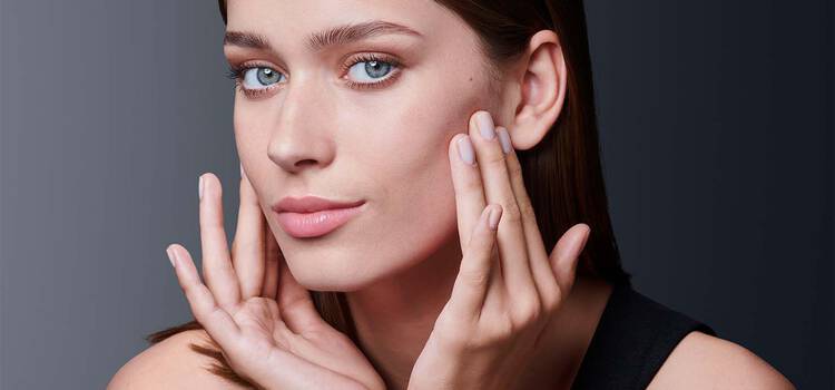 Eine Frau mit naturlichem Make-up und hellrosa Lippen beruhrt sanft ihr Gesicht mit beiden Handen und blickt direkt in die Kamera.