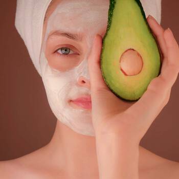 Frau hat eine Gesichtsmaske auf dem Gesicht und die Haare in ein Handtuch eingewickelt und halt eine halbierte Avocado vor dem Auge