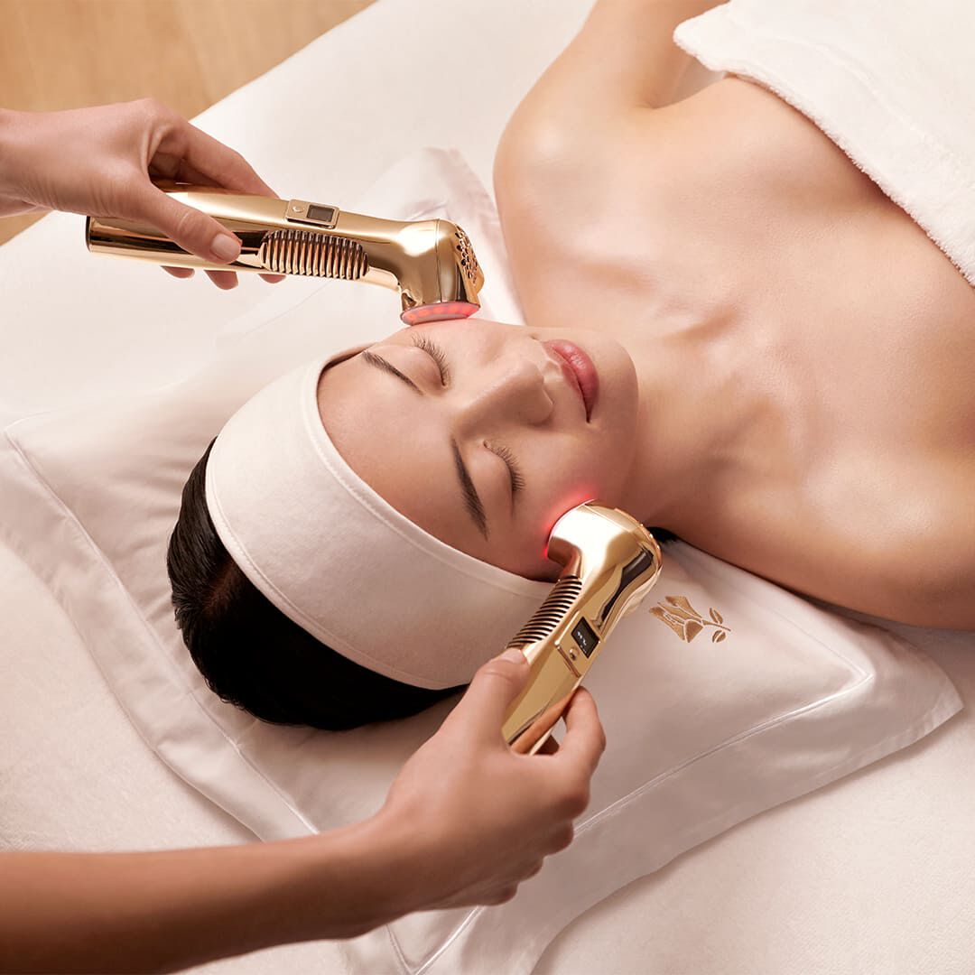 Eine liegende asiatische Frau erhalt eine professionelle Gesichtsbehandlung mit zwei goldfarbenen Lancome-Massagegeraten, die sanftes rotes Licht auf ihre Wangen abgeben, wahrend ihr Kopf auf einem weißen Kissen mit goldener Lancome-Rose ruht.