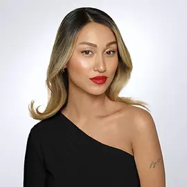 Eine Frau mit langen, glatten Haaren, die von dunkelbraun zu blonden Spitzen ubergehen. Sie tragt ein schulterfreies schwarzes Top und hat einen leuchtend roten Lippenstift aufgetragen. Der Hintergrund ist hell und neutral.